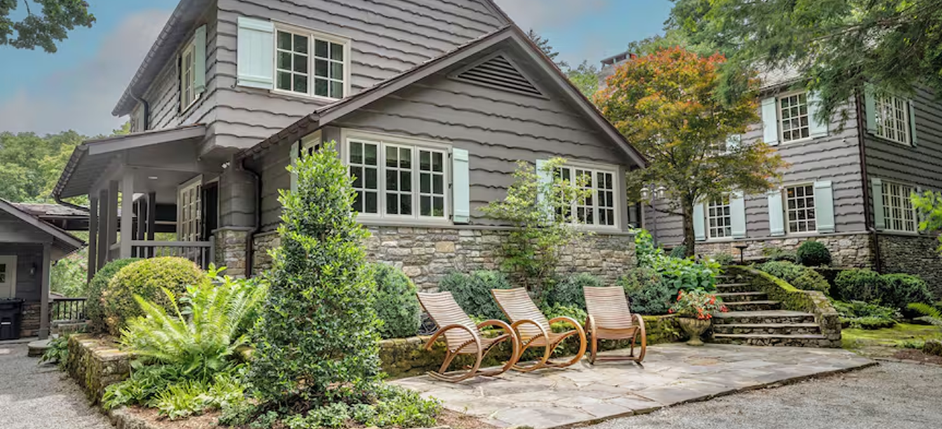 Cozy outdoor seating area with wooden chairs beside a gray house and greenery.