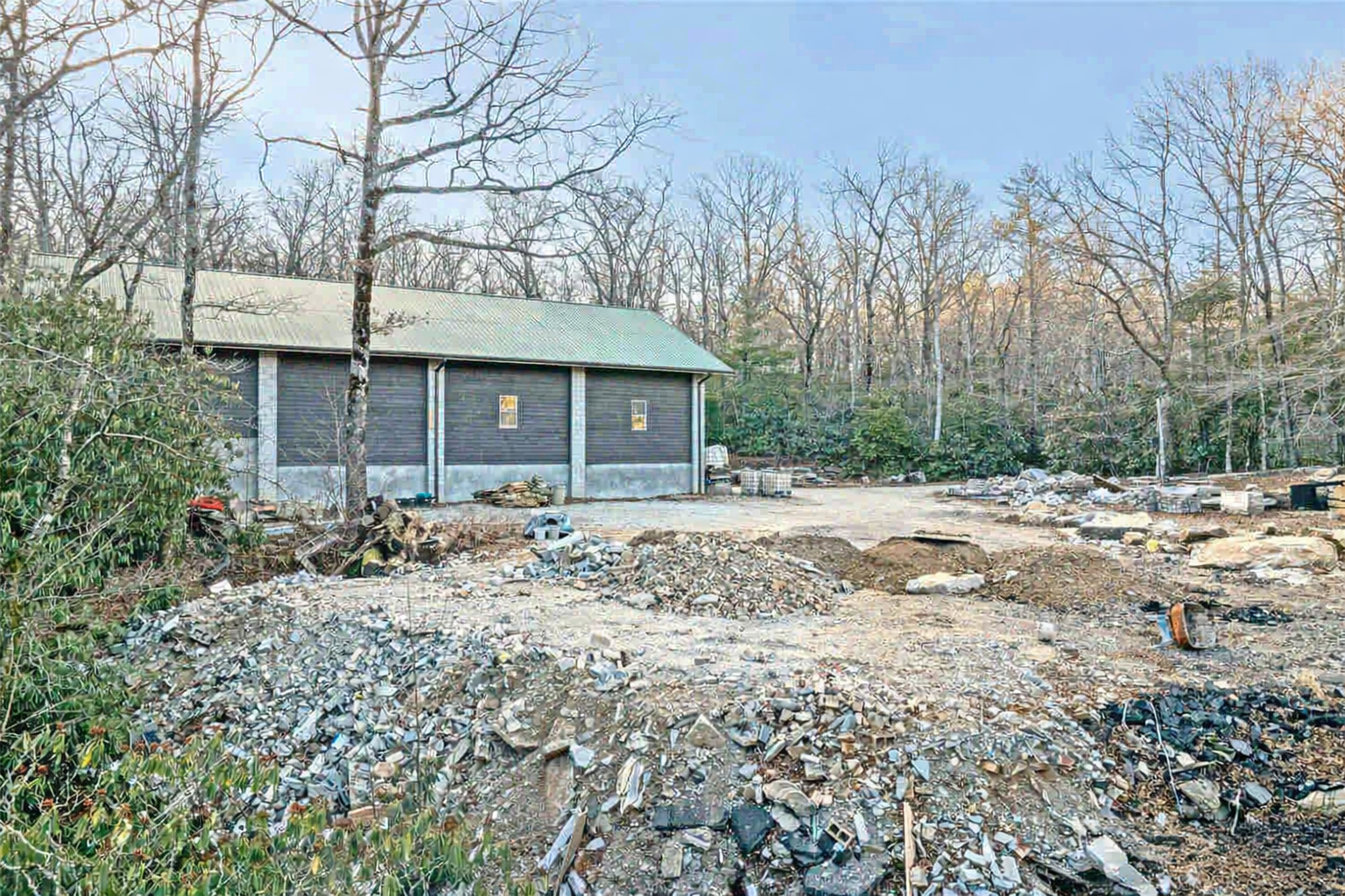 Stone foundation and cleared land next to a blue house in a wooded area.